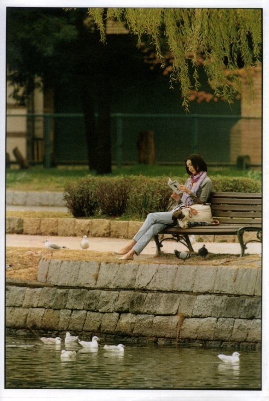 Woman seated on a park bench by water wearing spring womenswear from luxury brands including Moncler, Burberry Brit, and Gucci.