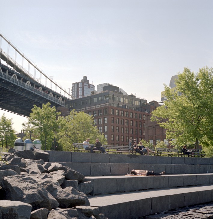 People relaxing on stone terraces at Brooklyn Bridge Park with Manhattan bridge and buildings in background.