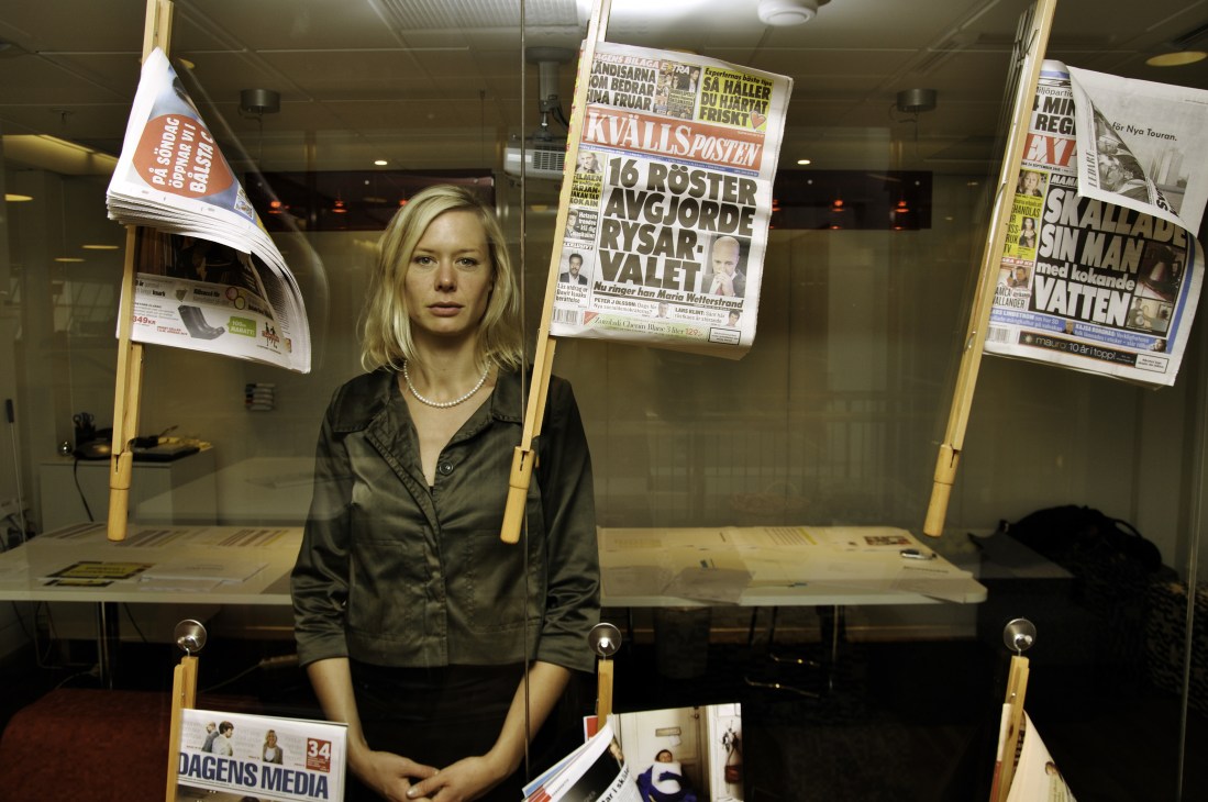 Woman in dark jacket standing in front of display of Swedish newspapers and publications