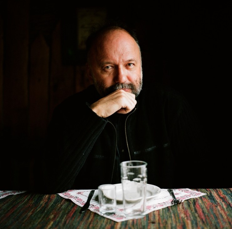 Ukrainian author Andrey Kurkov seated at a table, resting his chin on his hand with a contemplative expression.