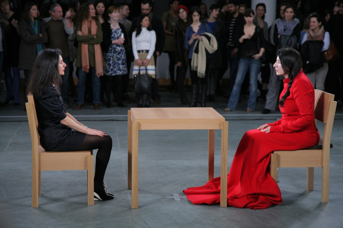 Marina Abramovic in red dress sitting across from a visitor in black at a wooden table during The Artist is Present performance.