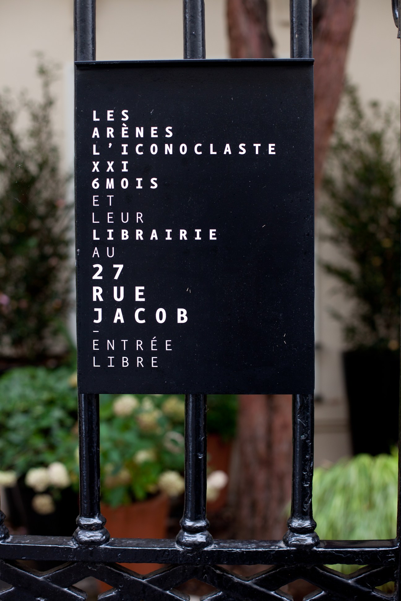 Black metal gate sign displaying Les Arènes publishing house and XXI magazine office location in Paris.