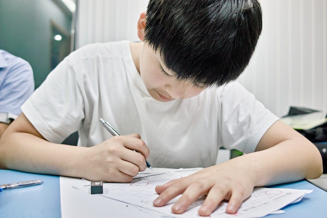 Young student in white shirt concentrating while writing on test paper at desk.