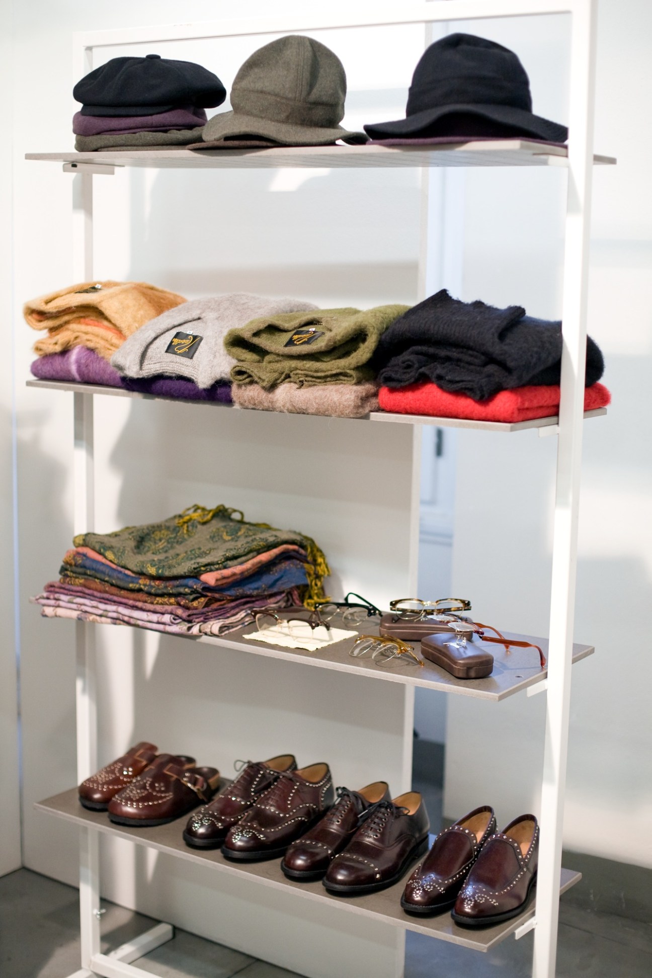 White shelving unit displaying Needles menswear collection including hats, knitwear, scarves, eyeglasses and studded brogues shoes.