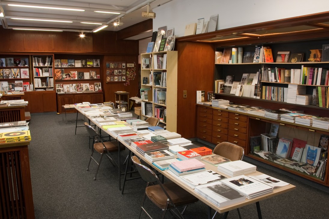 Interior of Motto bookstore and magazine shop in Berlin with wooden shelves, dark wood cabinetry, and a long table displaying publications and art books.