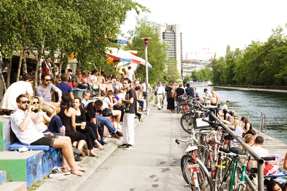 People relaxing by a canal in an urban park with bikes and trees.