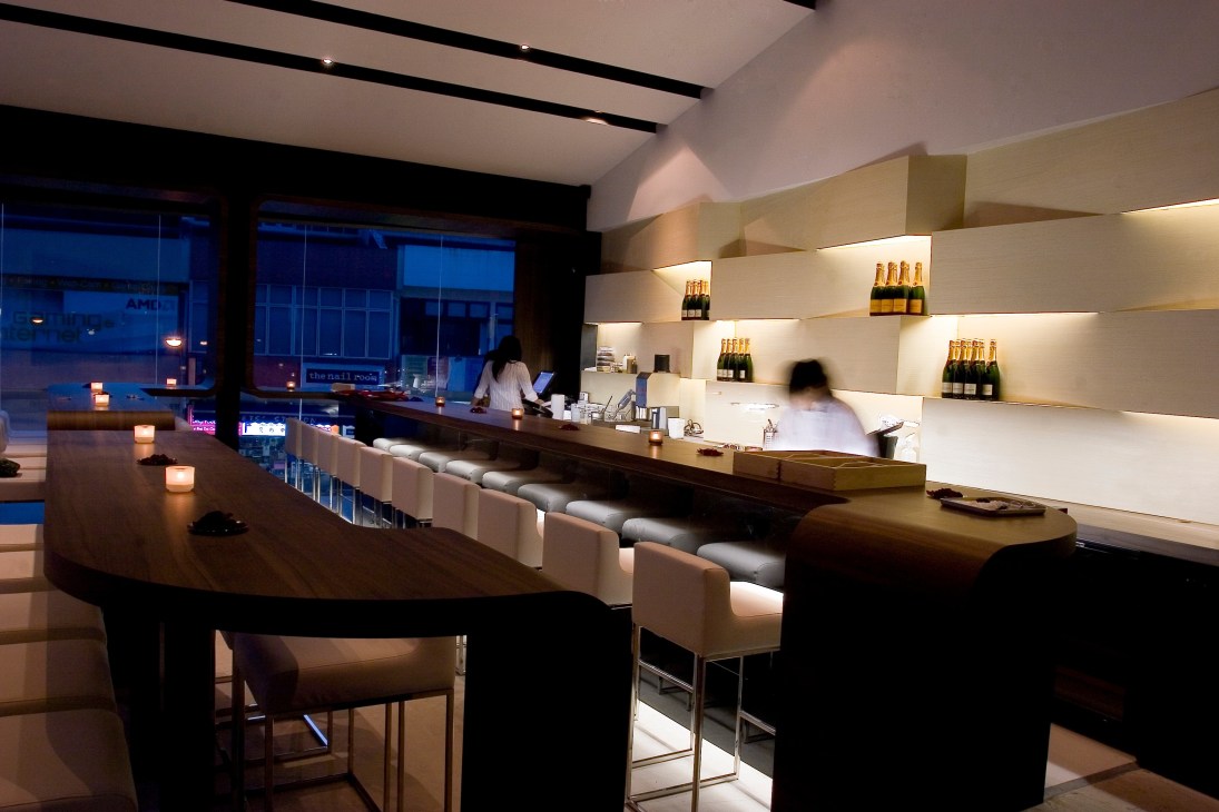 Modern dessert laboratory with illuminated shelving displaying bottles, dark wood counter seating, and floor-to-ceiling windows overlooking city at dusk.