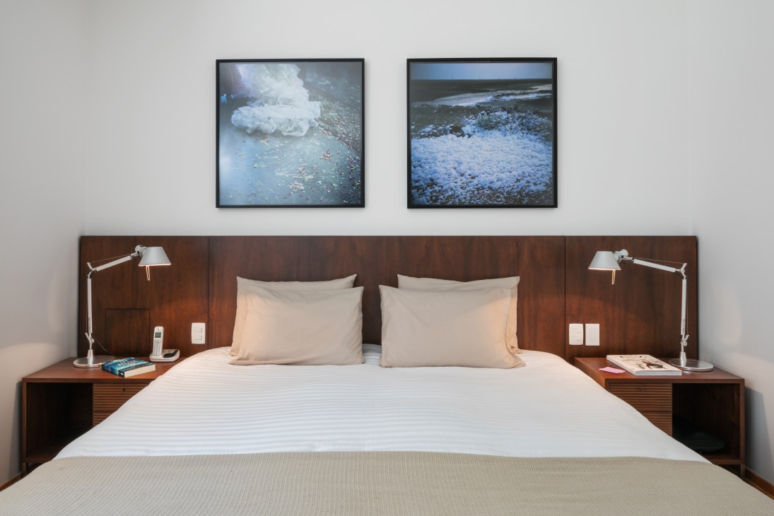 Modern bedroom with wooden headboard, white bedding, adjustable desk lamps, and two framed seascape photographs