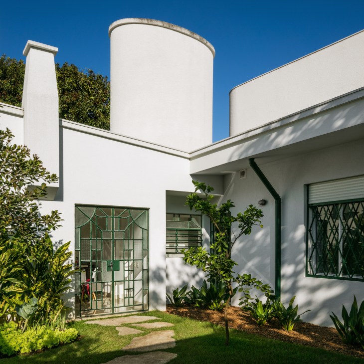 White modernist house with cylindrical tower, green iron window grilles and landscaped garden under blue sky