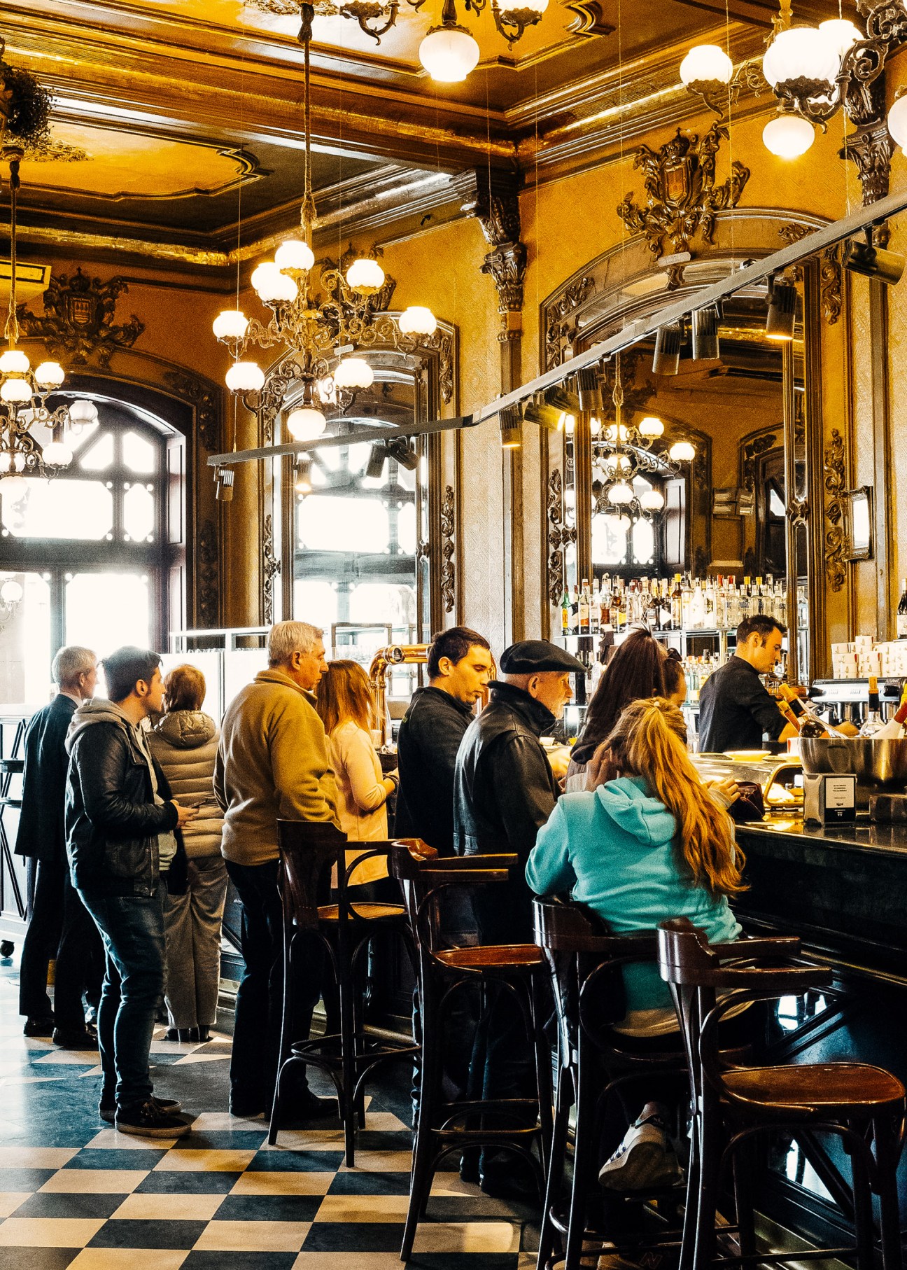 Ornate golden interior of Café Iruña in Pamplona with patrons at the bar beneath decorative chandeliers.