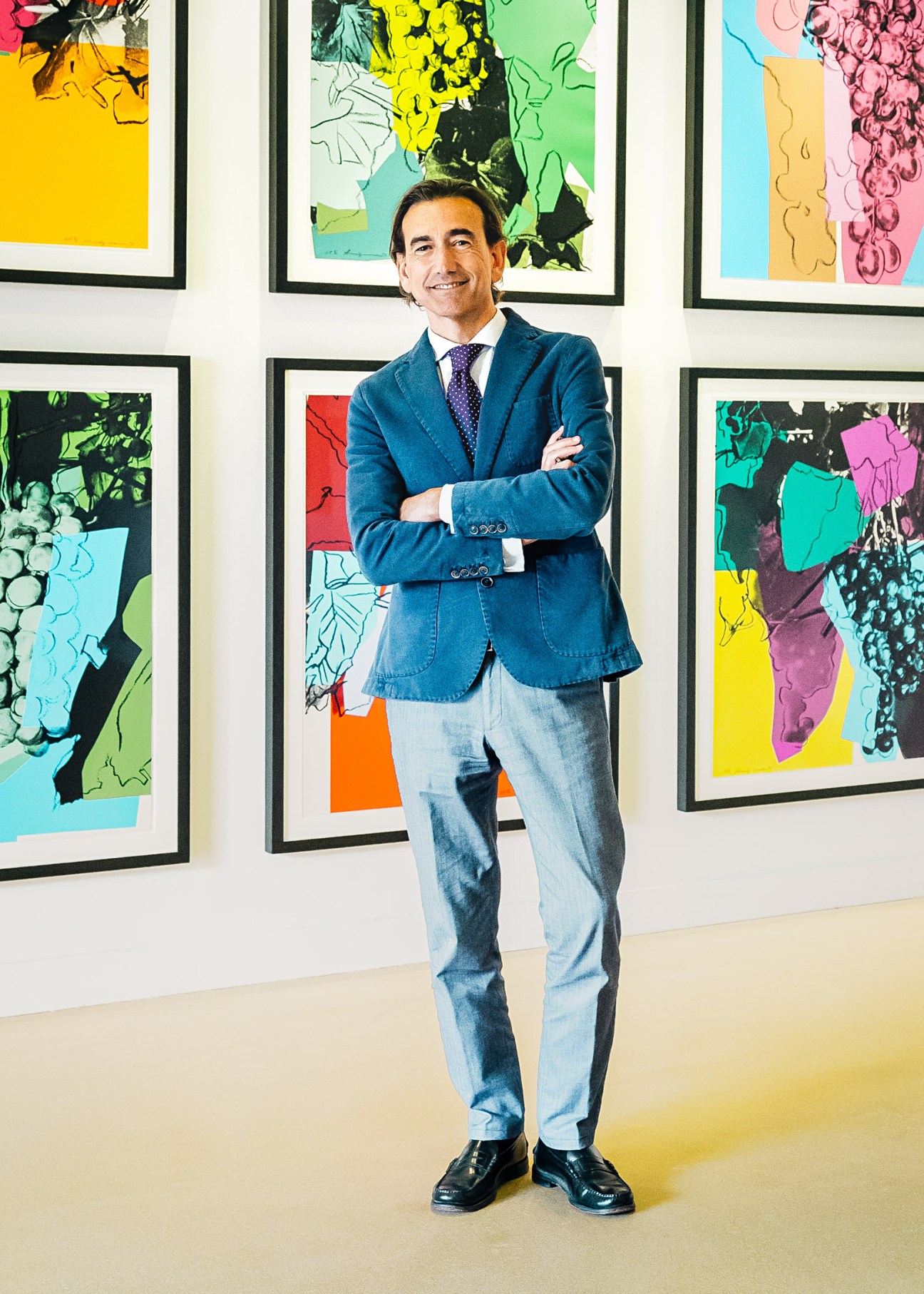 Man in blue blazer stands with arms crossed in front of colorful framed artworks.