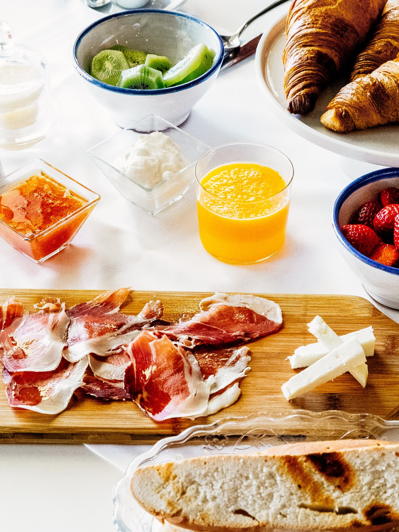 Breakfast spread with cured meats, croissants, fresh fruit, orange juice, and bread on a white table.