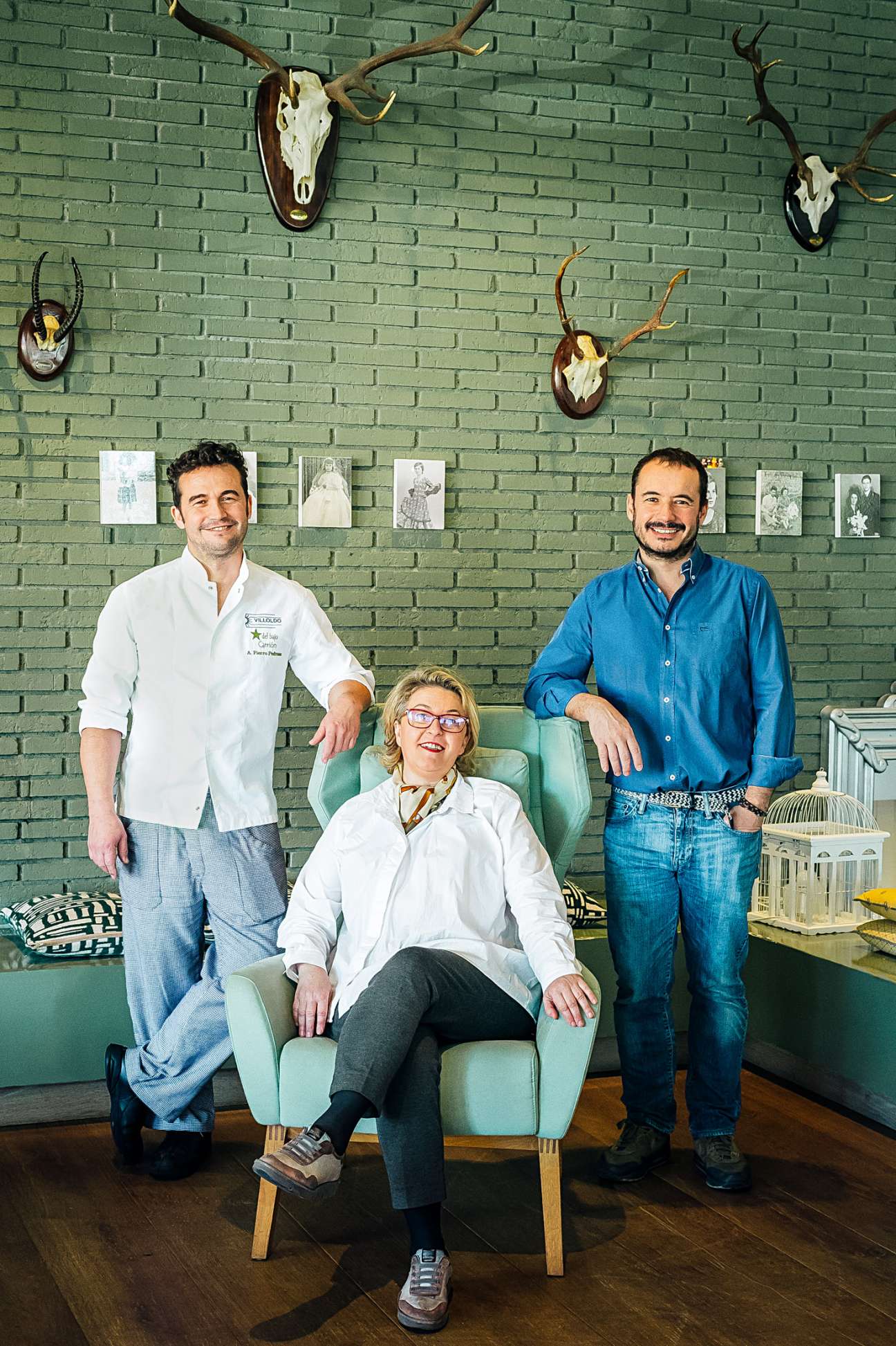 Family portrait at Estrella de Bajo Carrión showing three people posing in front of a green brick wall decorated with animal skulls and sketches.
