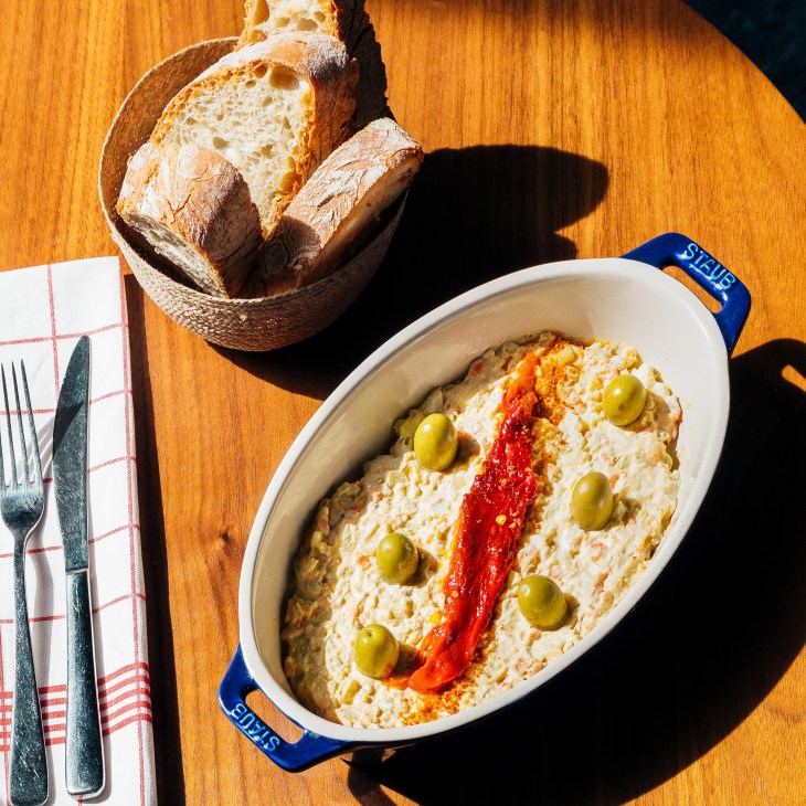 Ensaladilla Rusa in a blue Staub dish with bread and place setting on wooden table