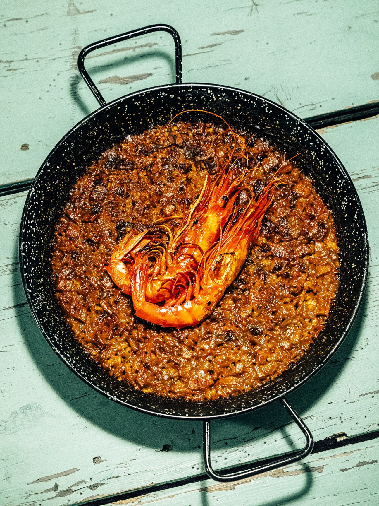 Seafood arroz in a black speckled paella pan with a cooked prawn on top, on a pale turquoise wooden surface.