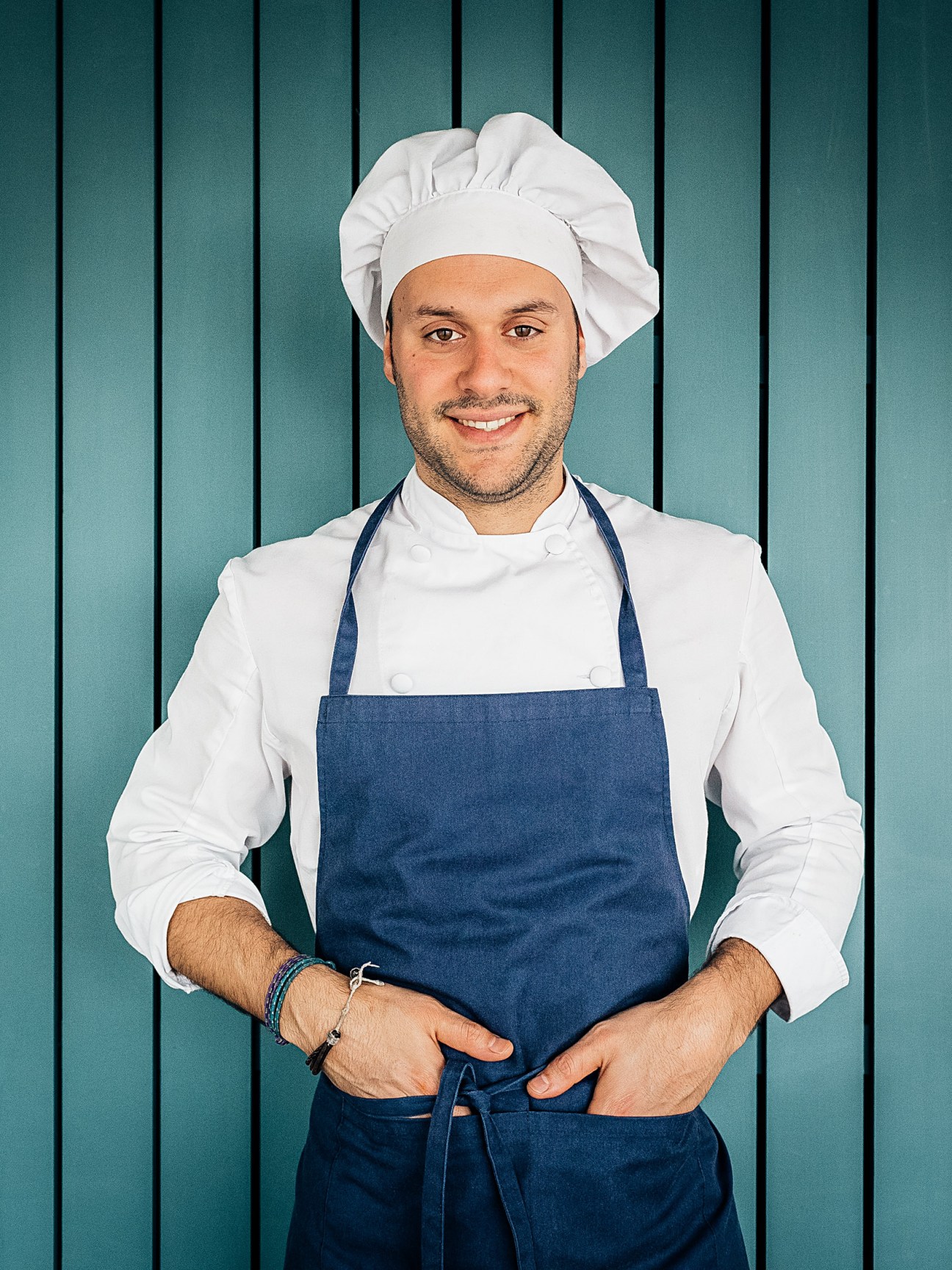 Chef wearing white chef's jacket, blue apron and white chef's hat against teal striped background.