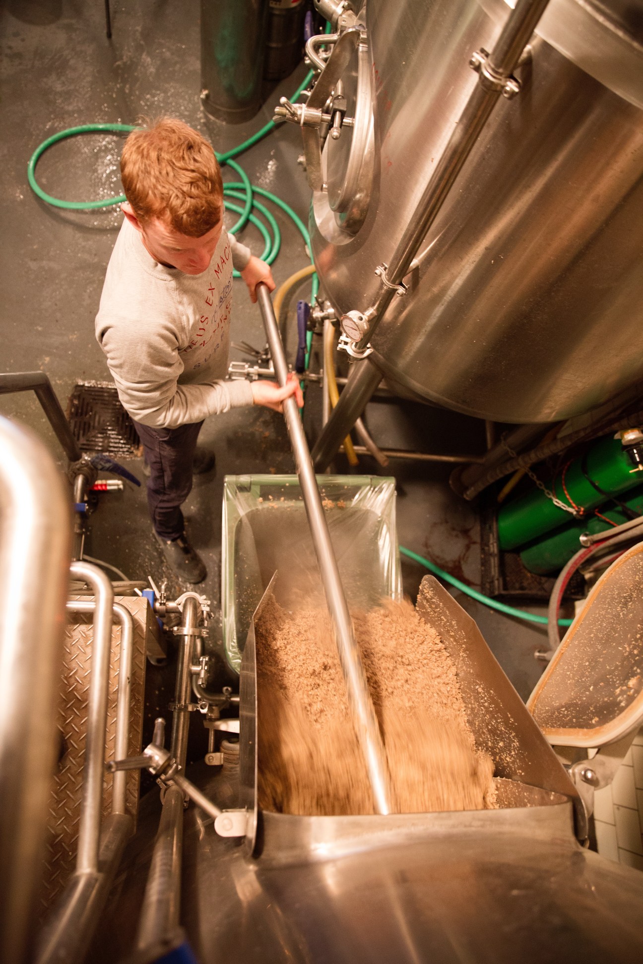 Brewer pouring malt grain into a large metal brewing vessel during beer production.