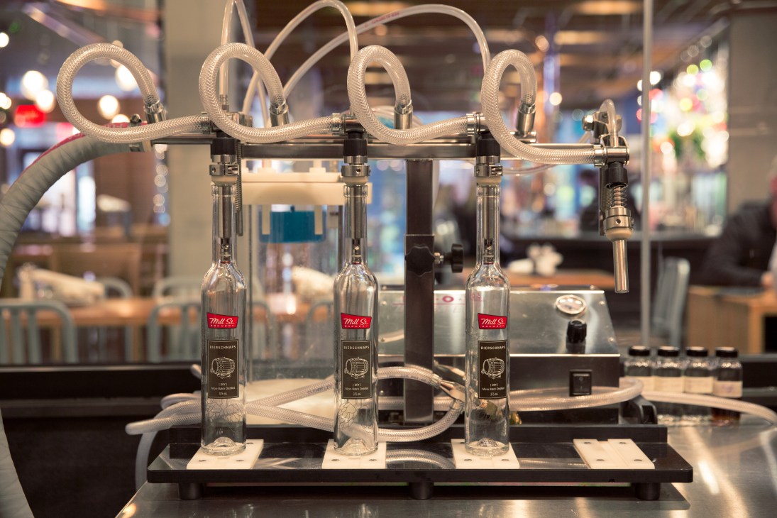 Mill St Bierschnaps bottles on a filling line with white tubing and nozzles.