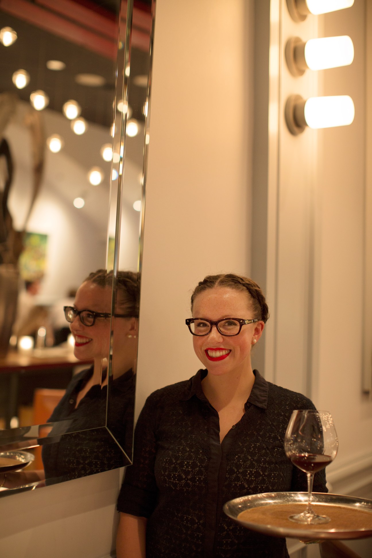 A smiling staff member in glasses and black shirt at Splendido restaurant's mirror-lined counter with illuminated vanity bulbs.