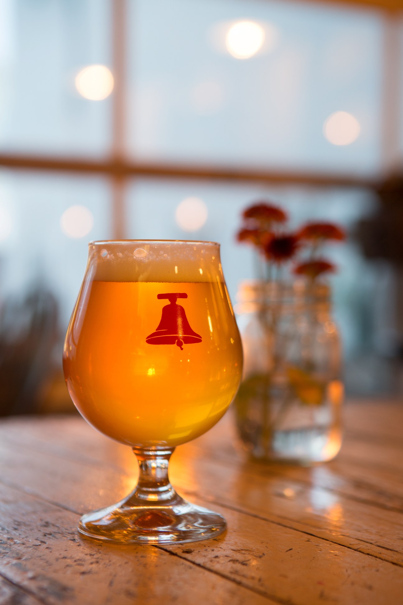 A snifter glass of amber-colored beer with a red bell logo, displayed on a wooden table with blurred windows and flowers in the background.