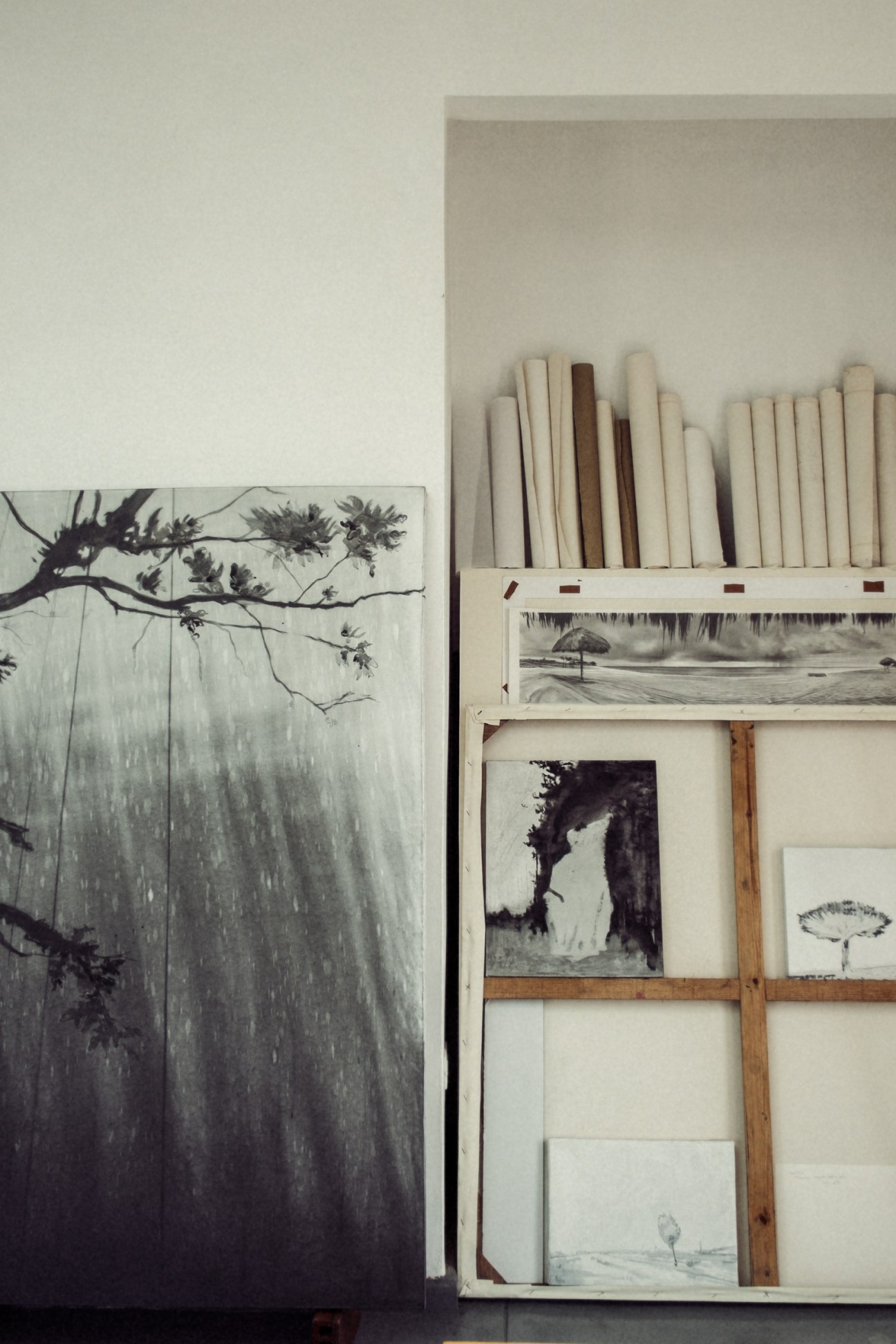 Large-format monochrome photograph of branches leaning against a storage unit with rolled papers and black-and-white photographs.