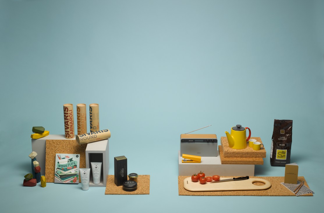 Styled product display featuring skincare, radio, stapler, notebooks, candles, coffee, chopping board and tea set against blue background.