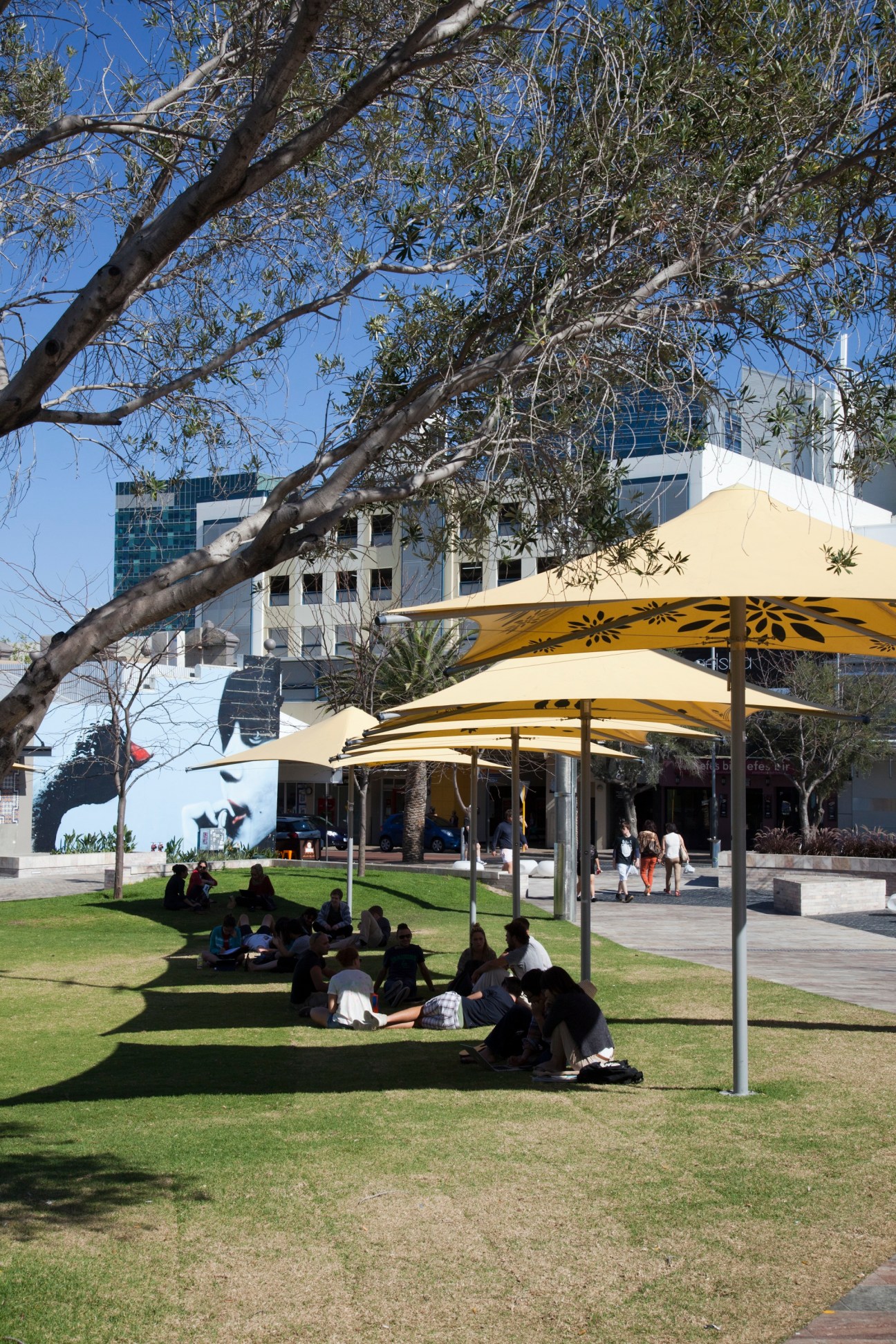 Groups of people sitting on grass under yellow shade sails in a public plaza with large trees and modern buildings.
