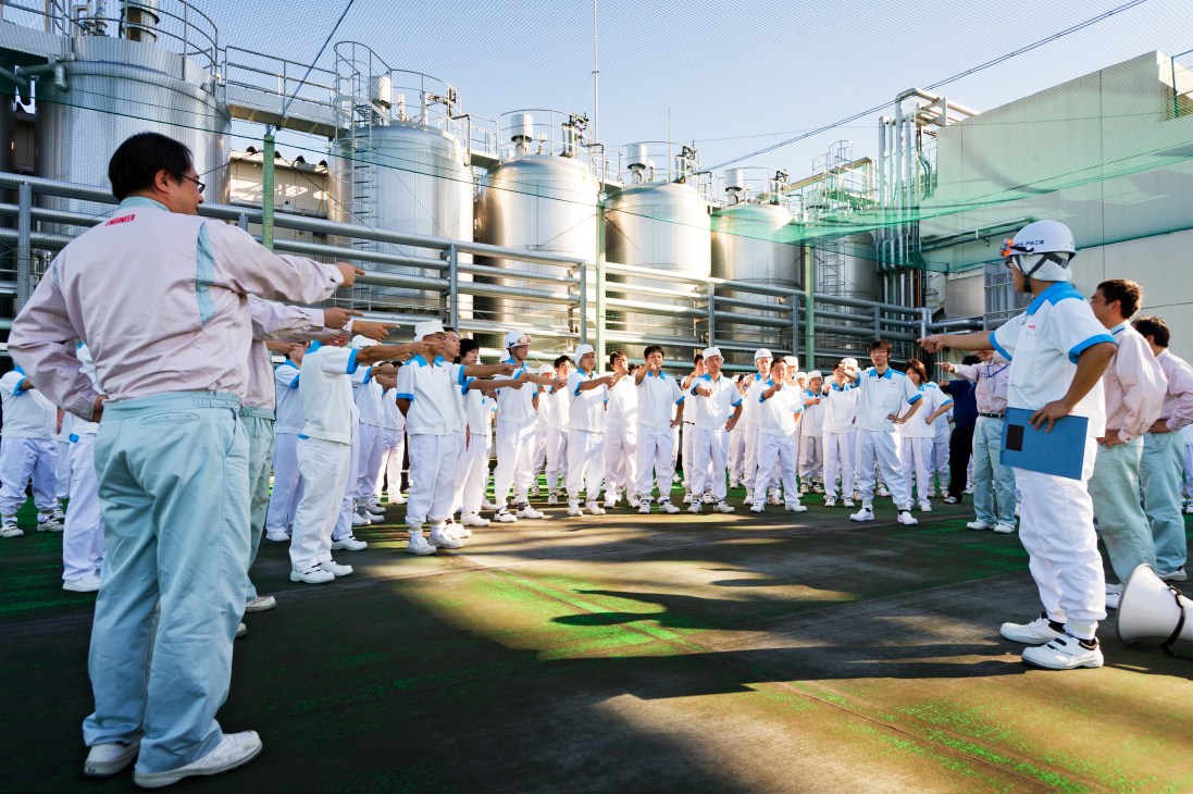 Workers in white uniforms perform synchronized rajio taiso exercises on a factory rooftop at Meiji's milk and yoghurt facility.