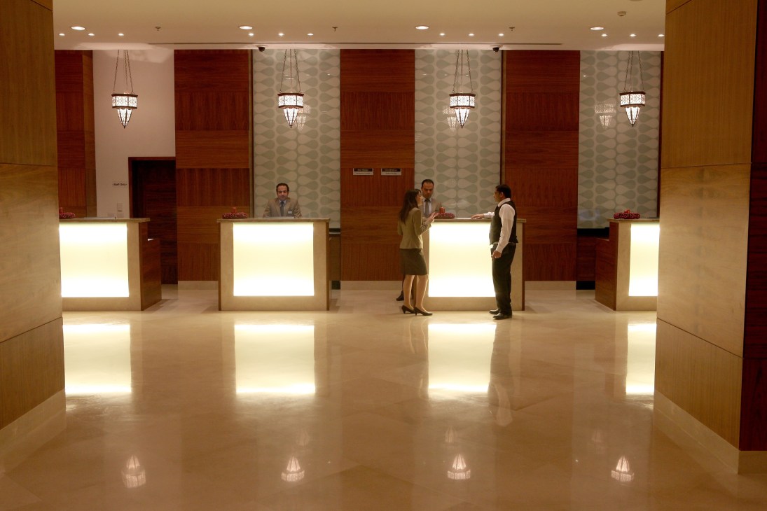 Modern hotel lobby with polished floors, illuminated reception desks, and guests conversing beneath decorative pendant lights.