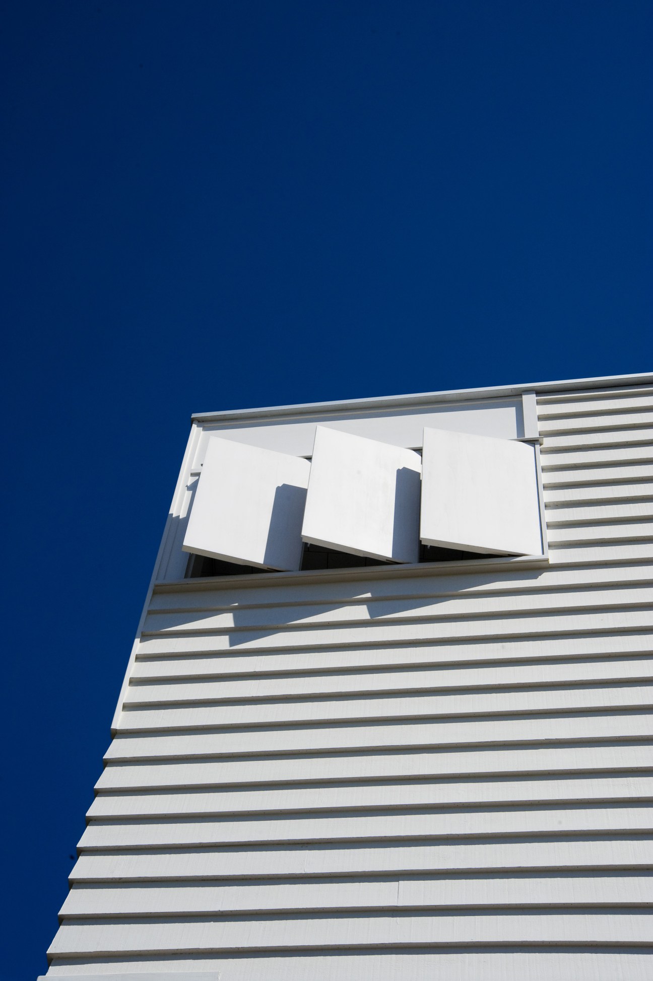Detail of white ventilation shutters and openings on a modern tower extension with horizontal weatherboard cladding against a blue sky.