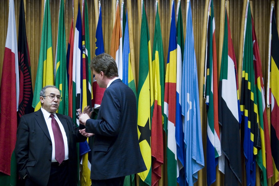 Two men in business suits converse in front of a display of African nation flags at the AU headquarters.