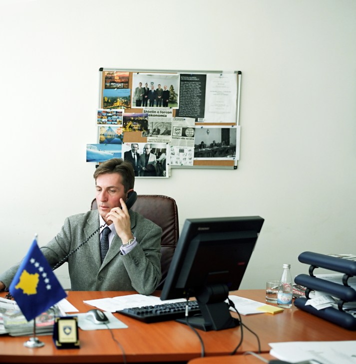 Lulzim Mjeku sits at his desk in the Ministry of Foreign Affairs office, speaking on a telephone with the Kosovo flag visible.