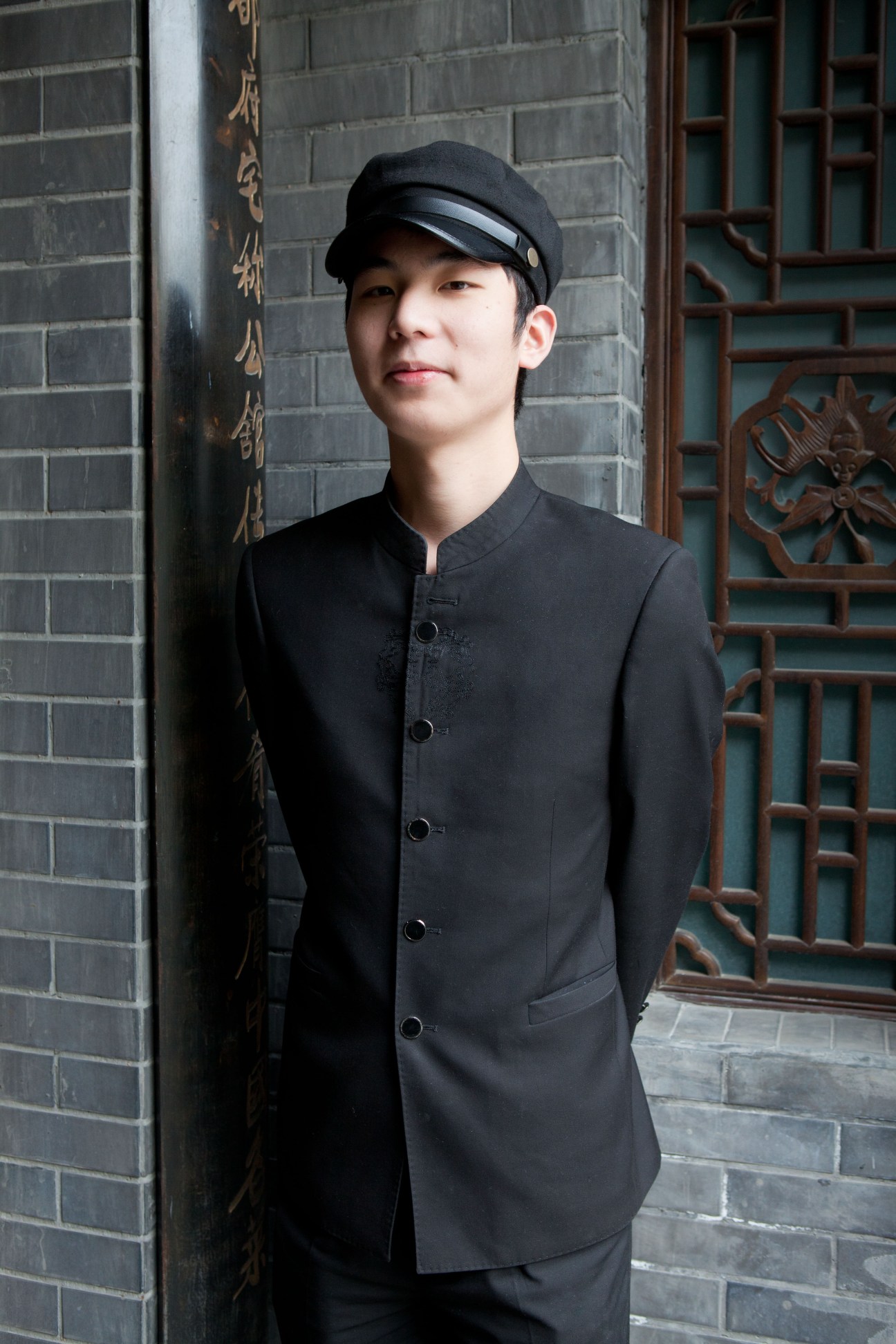 Young man wearing a black Mandarin-collar shirt and cap stands in front of traditional brick and wooden architecture in Kuanzhai Alleys.