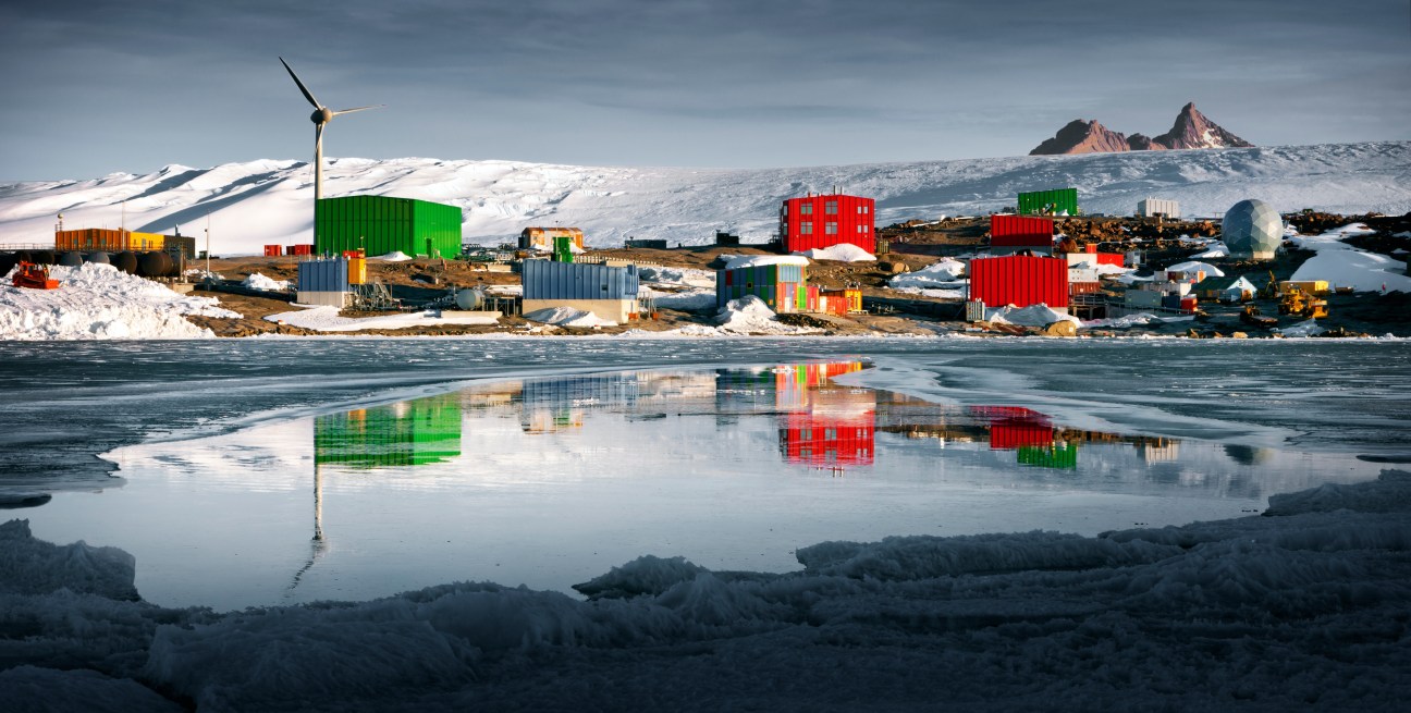 Colorful shipping containers and buildings at an Antarctic research station reflected in icy water, with snow-covered mountains in background.