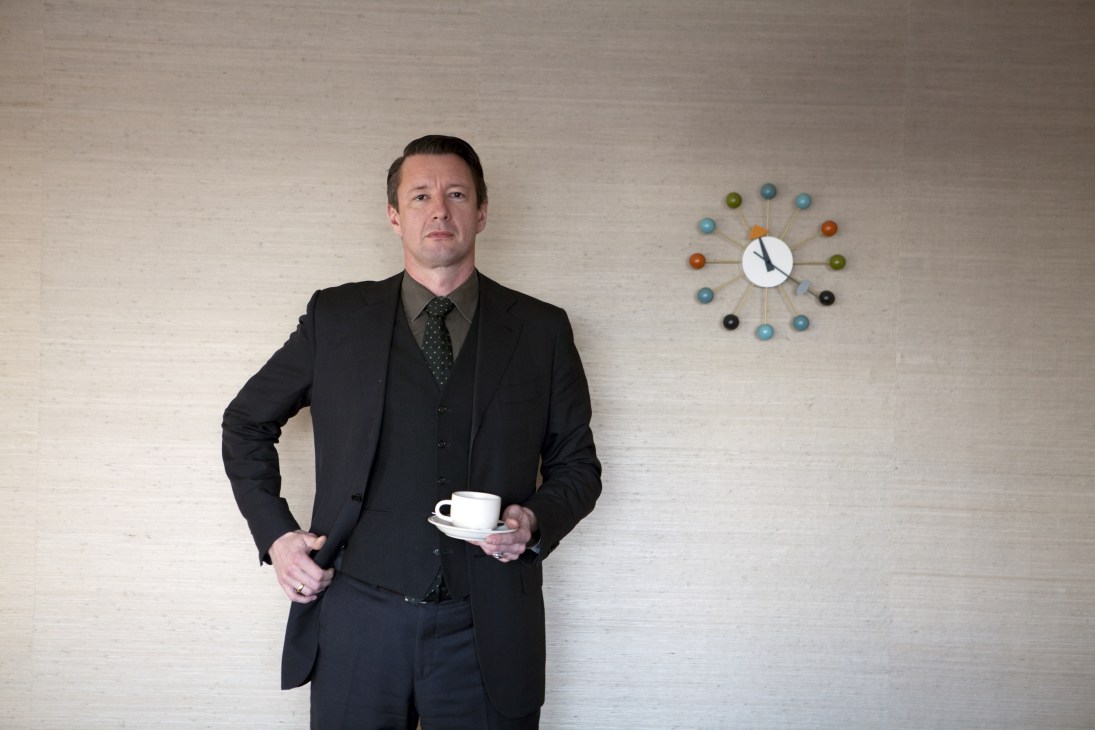 Man in dark suit holding white coffee cup stands against white wall with colorful clock.