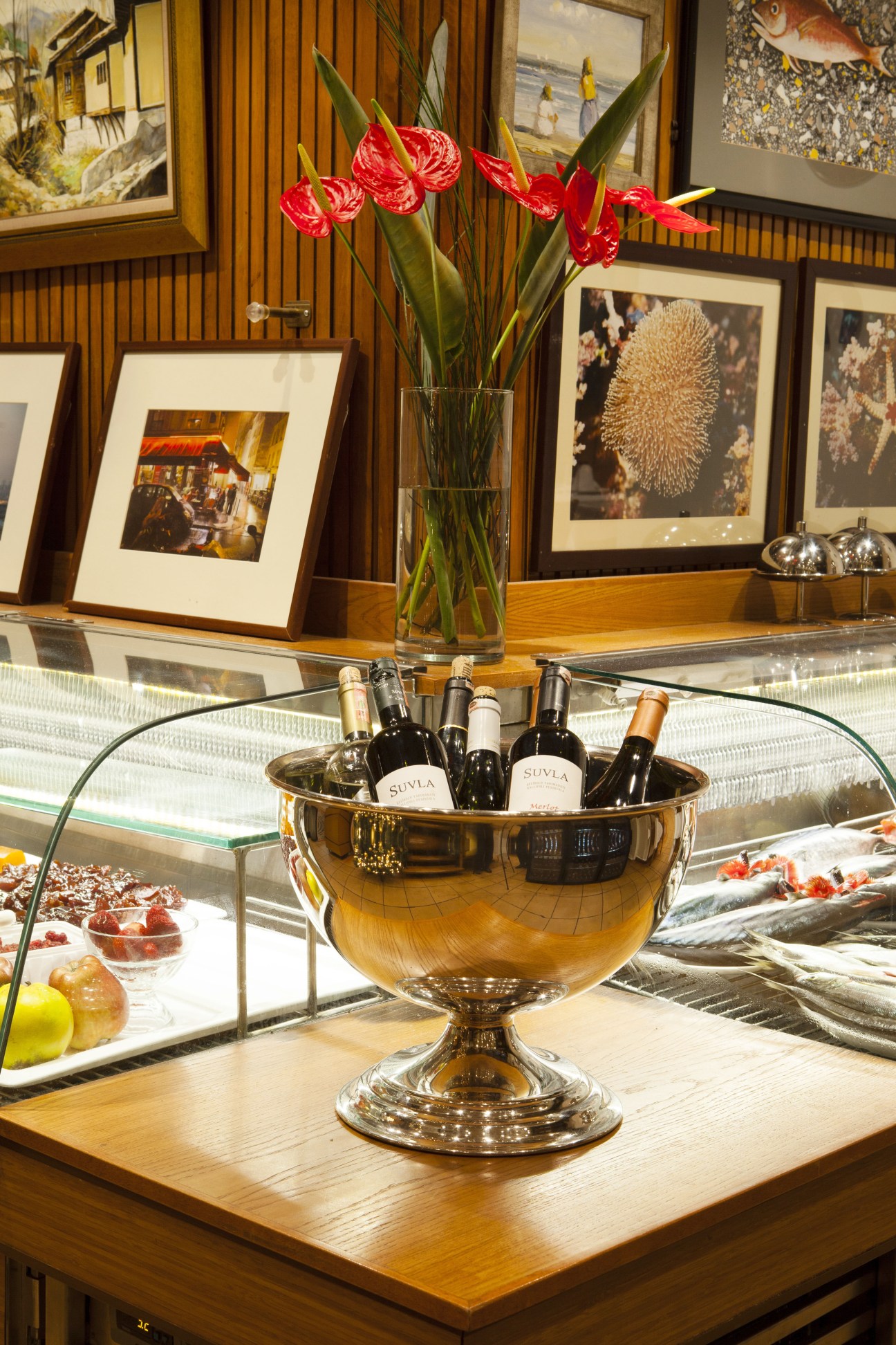 Wine bottles in a metal bowl with red flowers and framed photographs on wooden paneled wall at Kiyi restaurant.