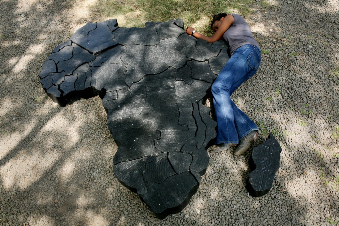Artist Ato Abe lies on ground next to a wooden jigsaw-like map of Africa.