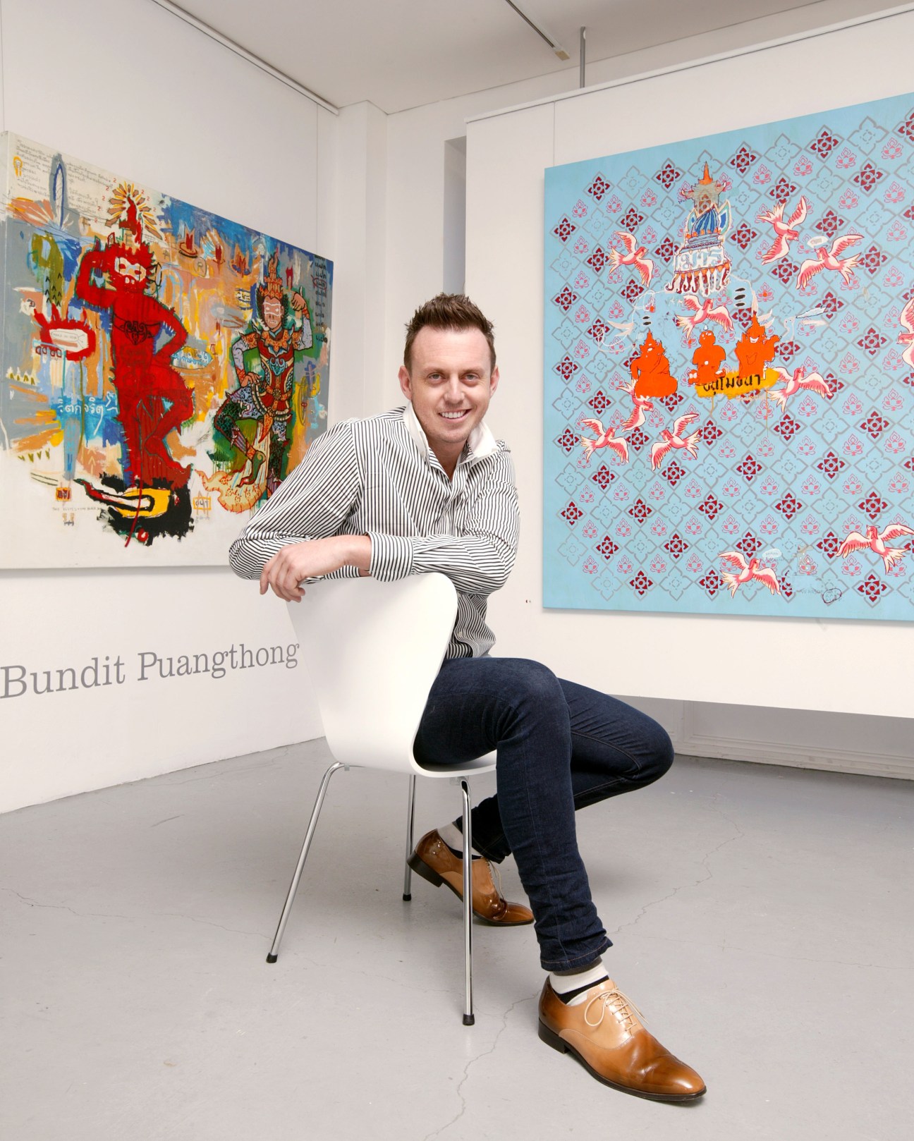 Iain Dawson poses in his gallery surrounded by colorful contemporary artworks including Thai-inspired paintings.