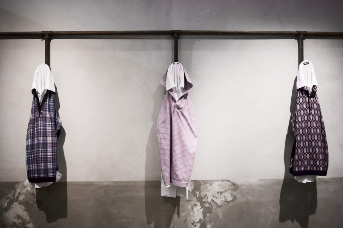 Three Drumohr knit jumpers displayed on hangers with white hoods, featuring tartan and argyle patterns in navy and purple tones.