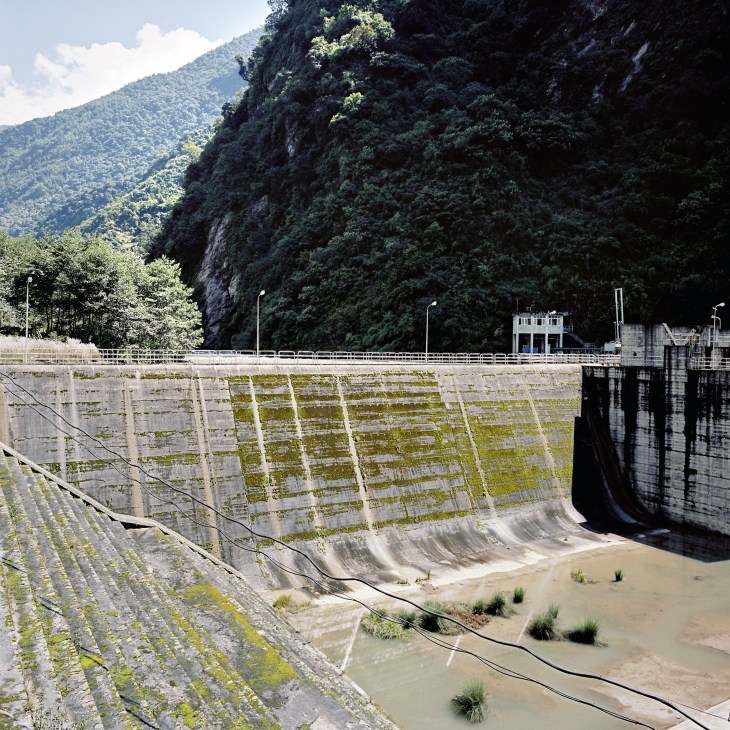 A concrete dam with moss-covered surface in a forested mountain valley with river below.