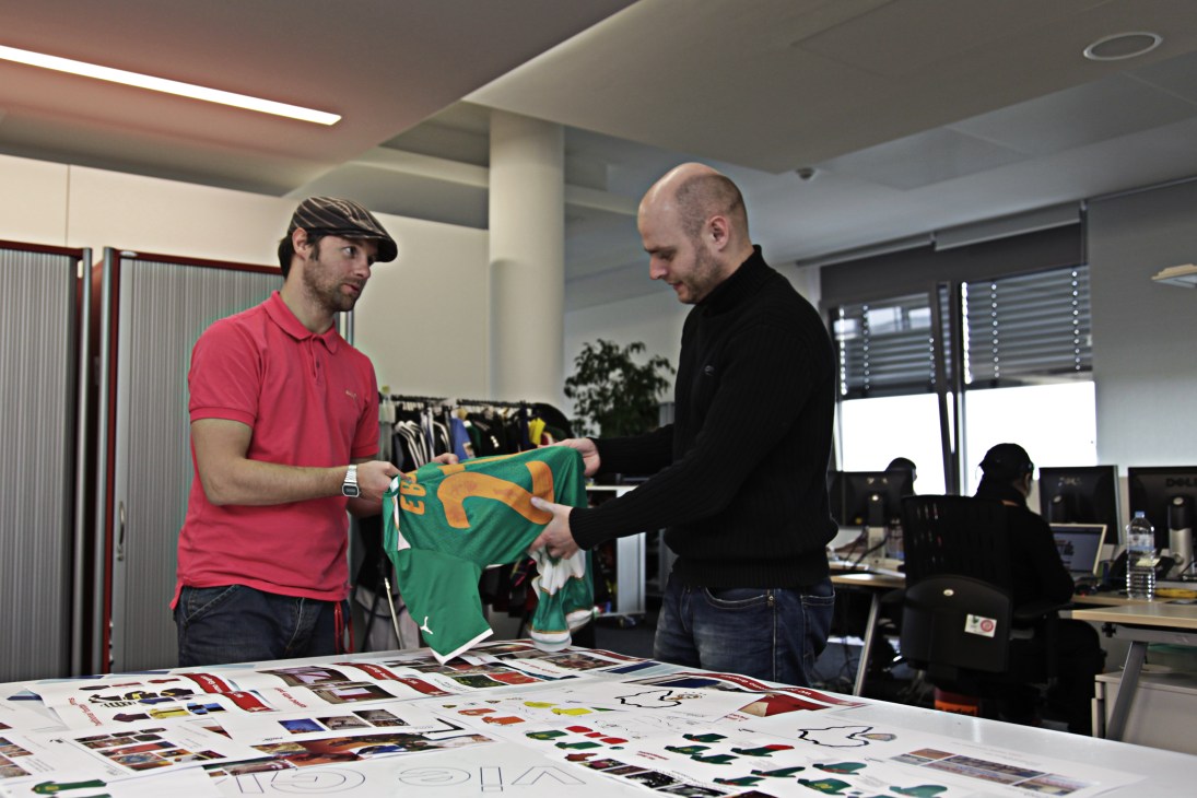 Two men examine a green football kit at a design table covered with sketches and samples at Puma headquarters.