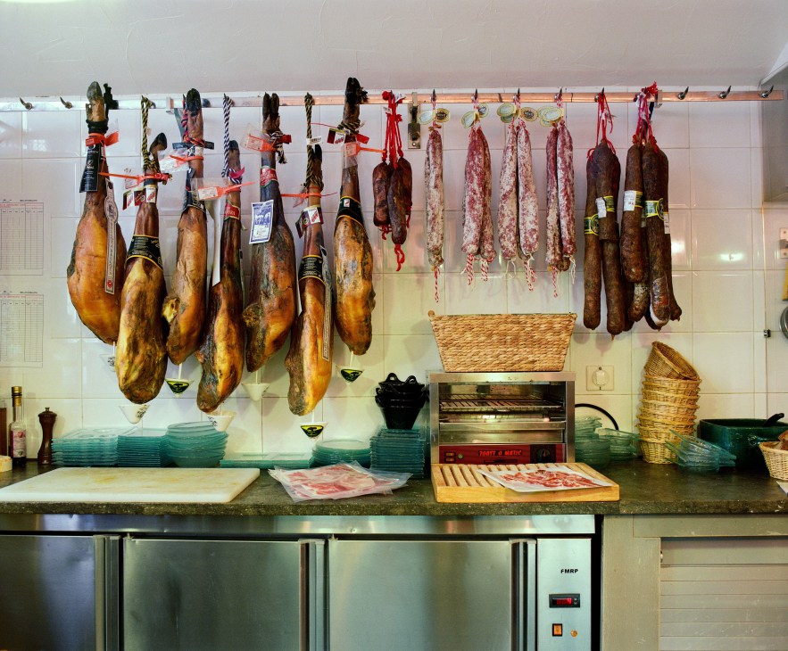 Cured Spanish jamón Ibérico and various charcuterie hanging from hooks on a kitchen wall at Bellota-Bellota.