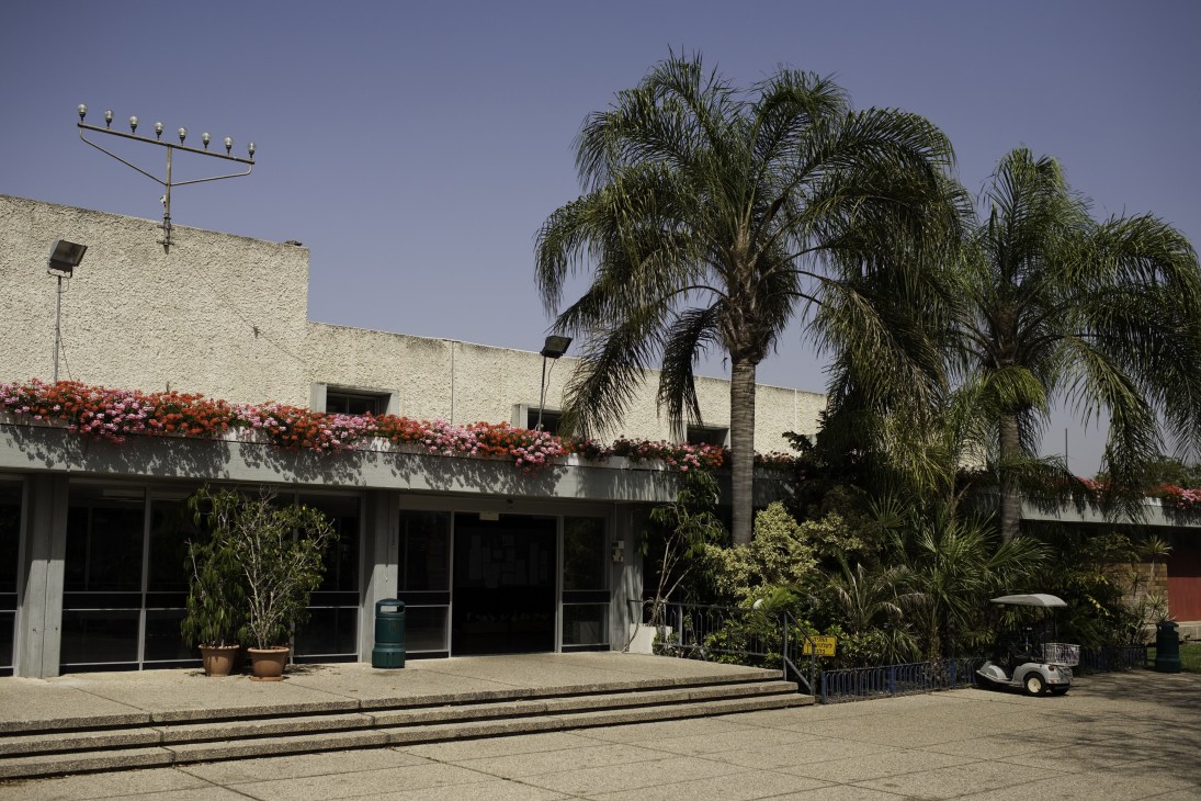 Main building of Maabarot kibbutz with modernist architecture, palm trees and flowering vines.
