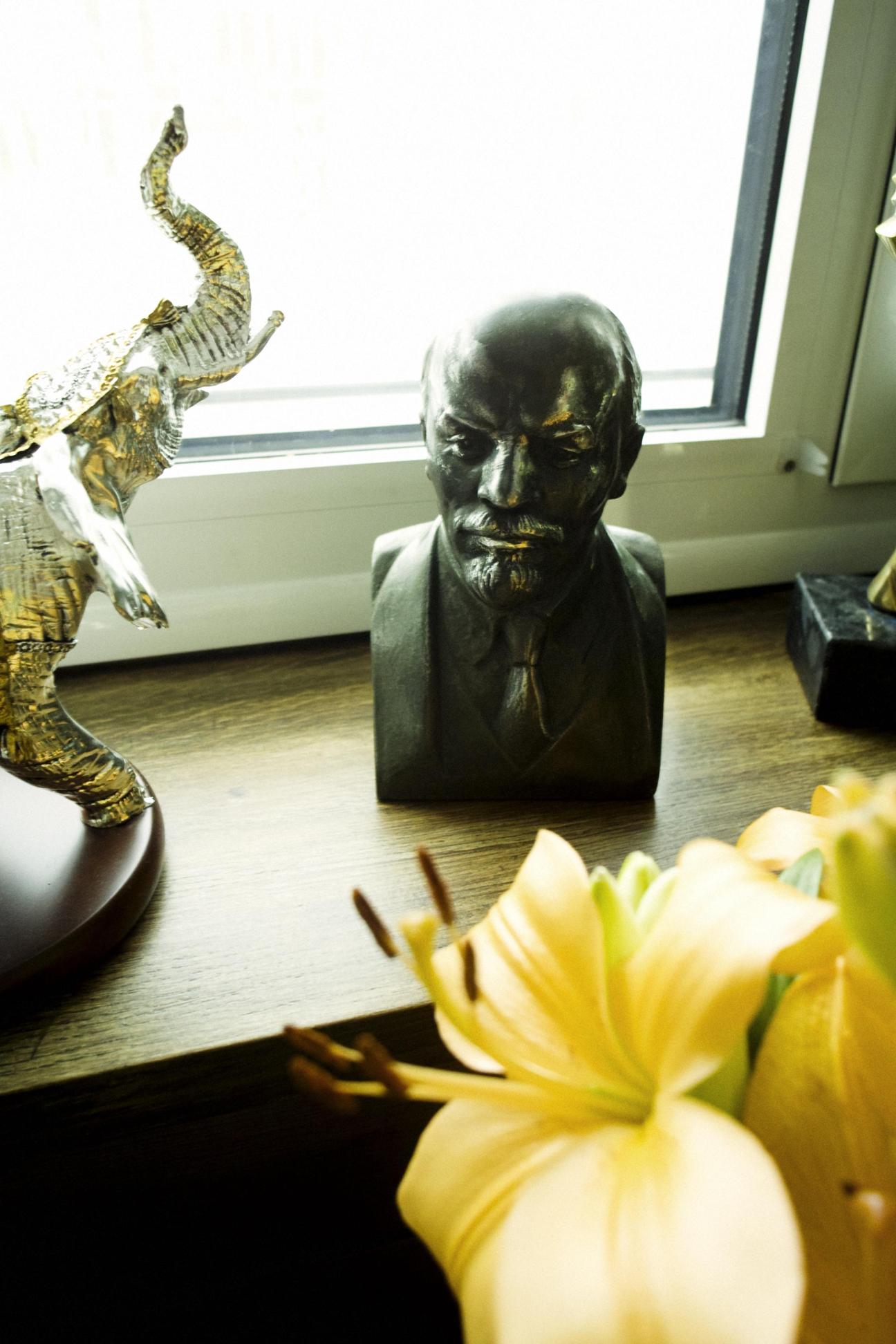 A dark bust sculpture of Lenin on a wooden shelf surrounded by decorative objects and yellow flowers.
