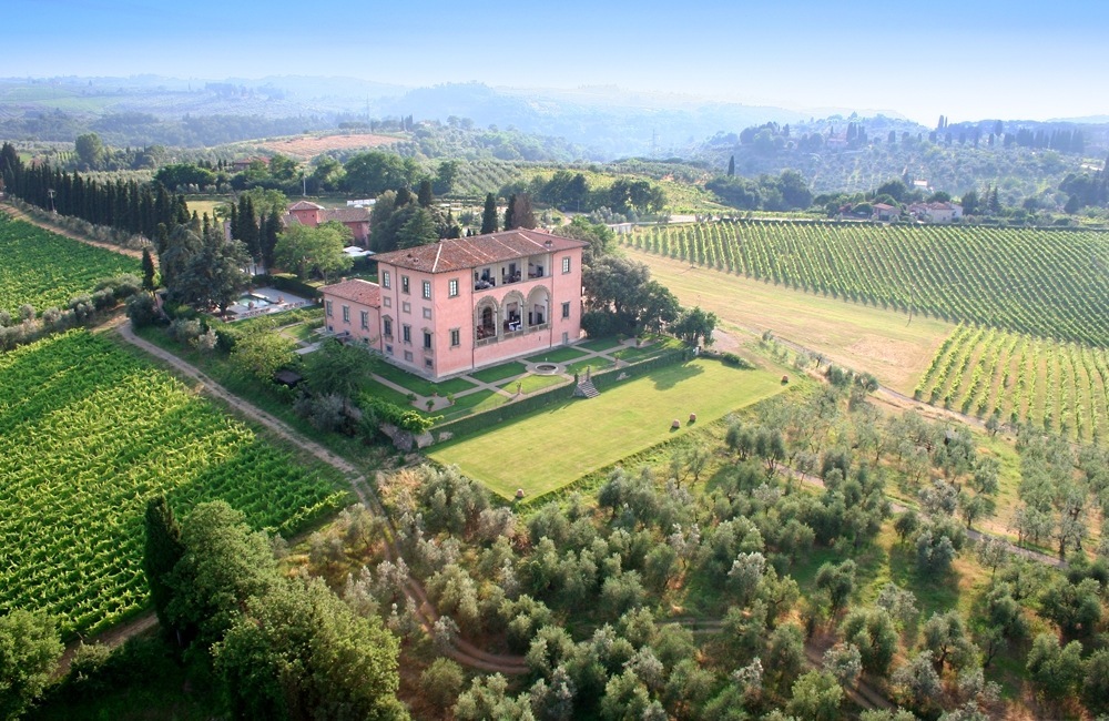 Aerial view of a pink Tuscan palazzo surrounded by vineyard-covered hills and cypress trees.