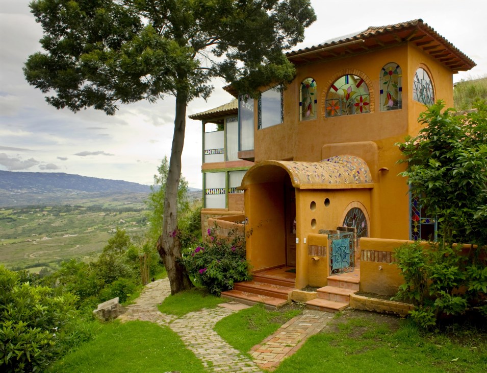 Yellow ochre-colored country house with decorative windows and arched entrance overlooking green valley landscape.