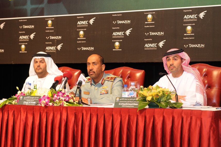 Three men seated at a press conference table with microphones and floral arrangements at IDEX 2009.