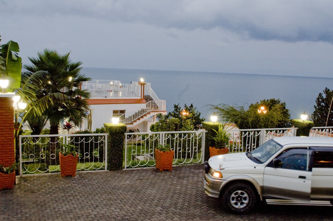 Upmarket lakeside residence with decorative gates, palm trees, and white SUV parked on brick driveway overlooking Lake Kivu.