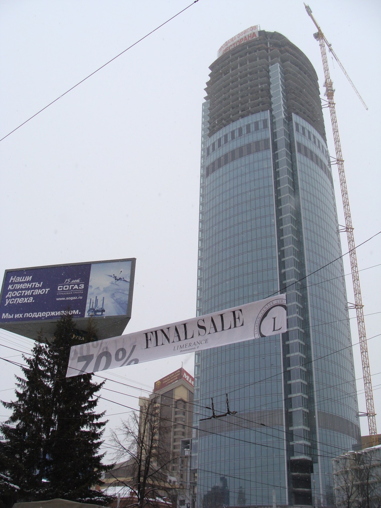 A 54-storey glass and concrete skyscraper under construction with a construction crane beside it, with a luxury goods store displaying a 70% final sale sign in the foreground.