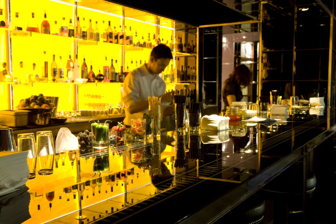 Brightly lit yellow bar at Rojo y Blanca with backlit shelving, bartender, and dining area.